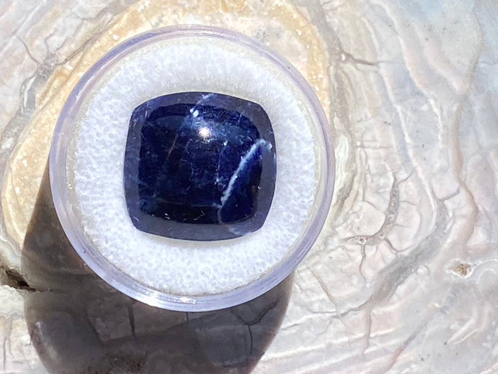 Blue gemstone in a clear plastic container on a textured white background