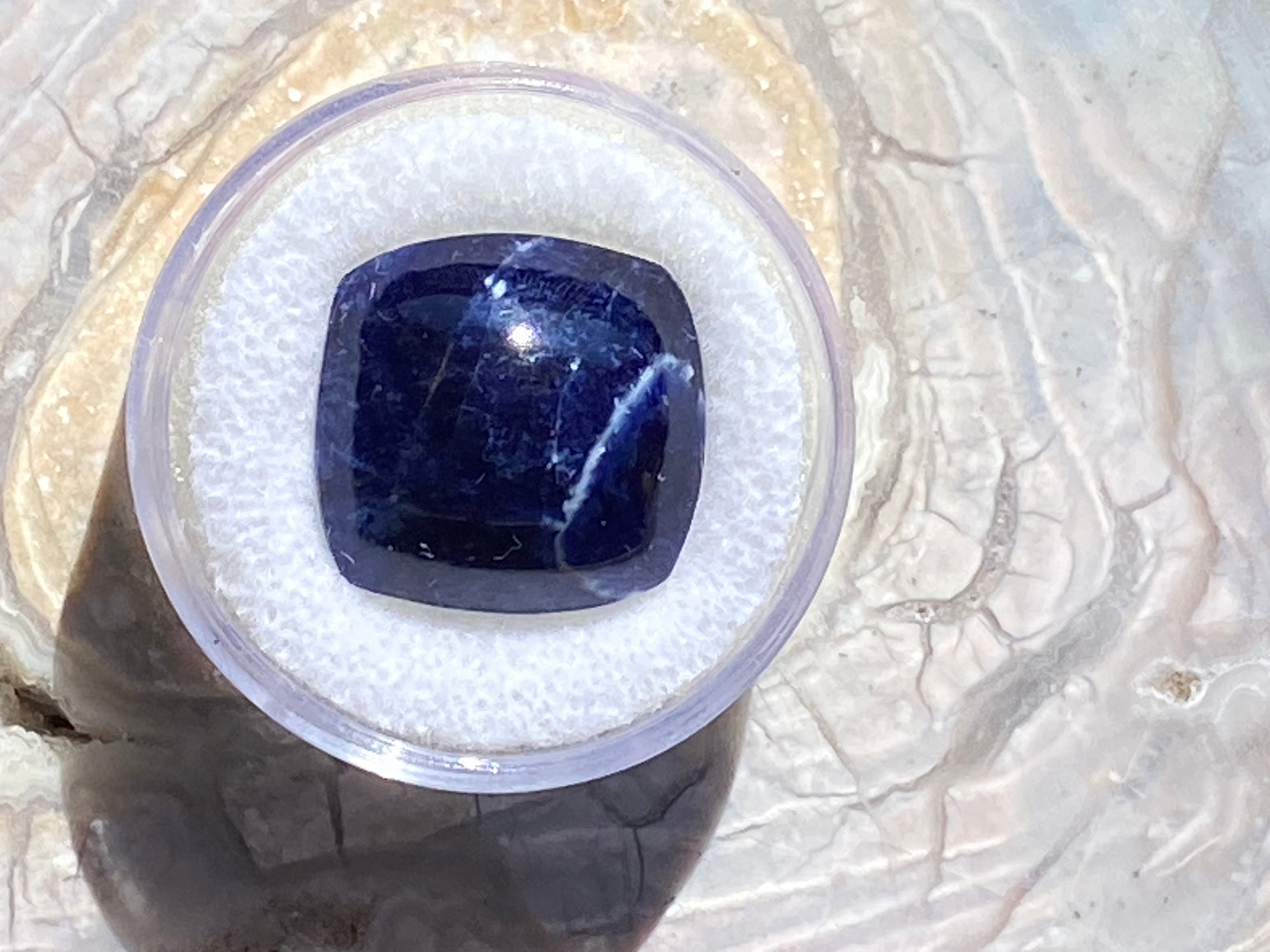Blue gemstone in a clear plastic container on a textured white background