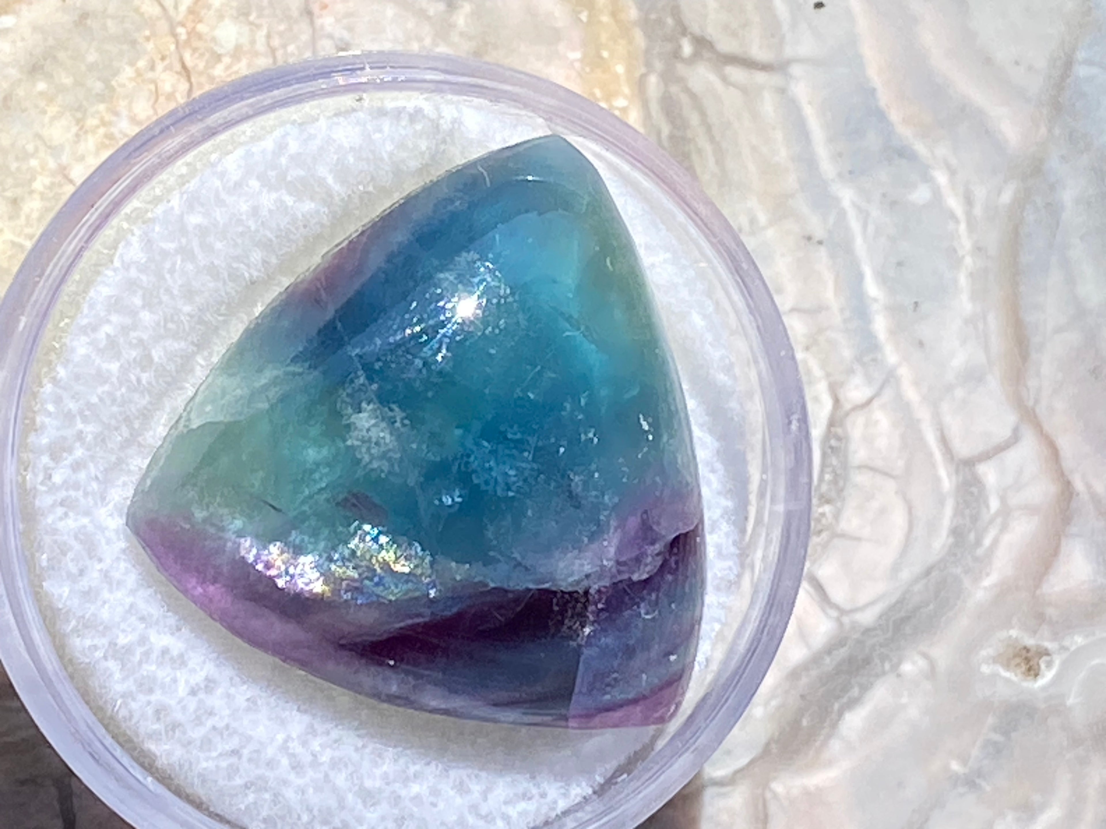 Triangular gemstone with purple and blue hues in a clear container on a textured white background