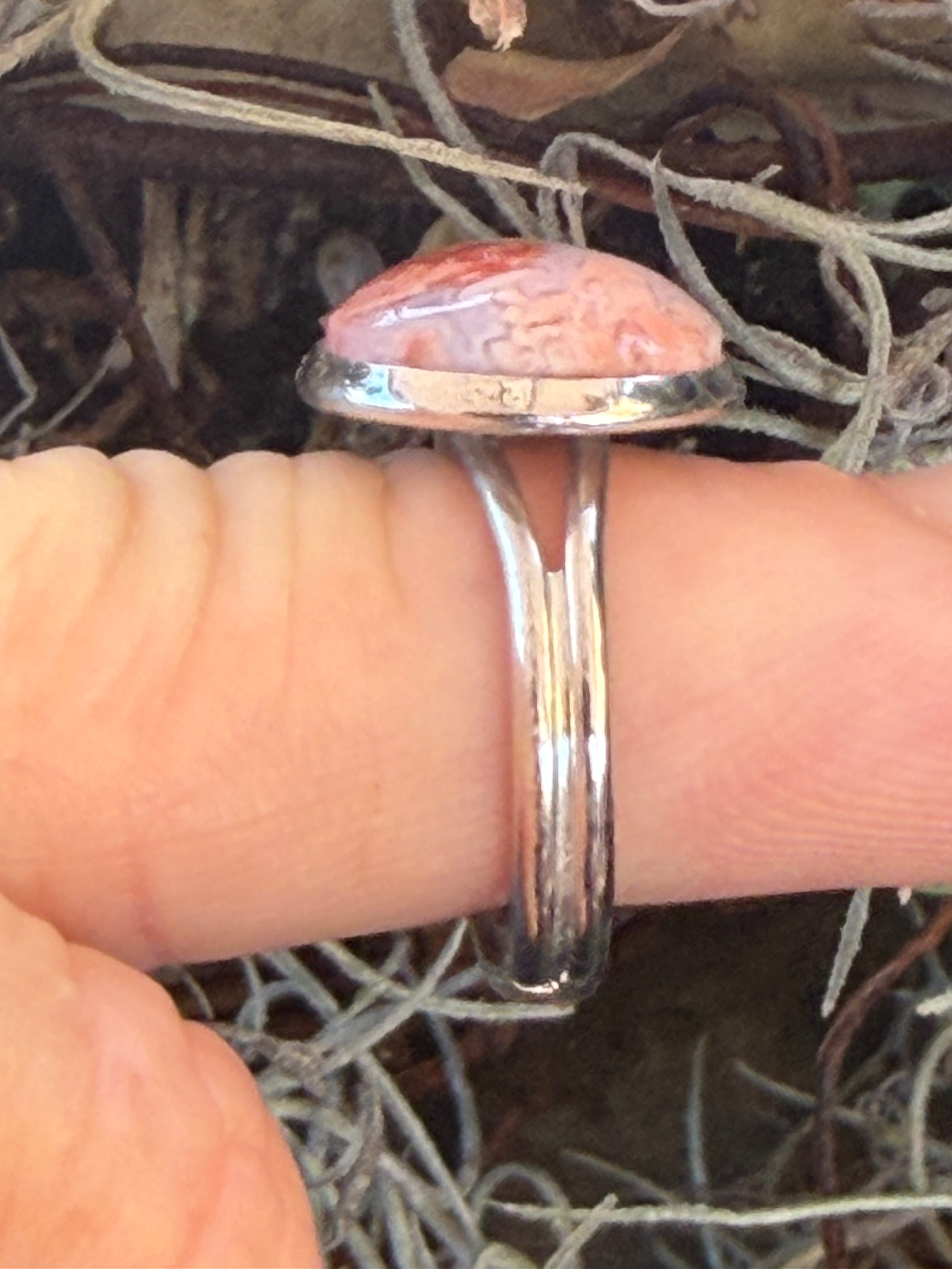 A silver band ring with a large, irregularly shaped pinkish-orange stone, shown worn on a finger against a background of dry grass and soil.