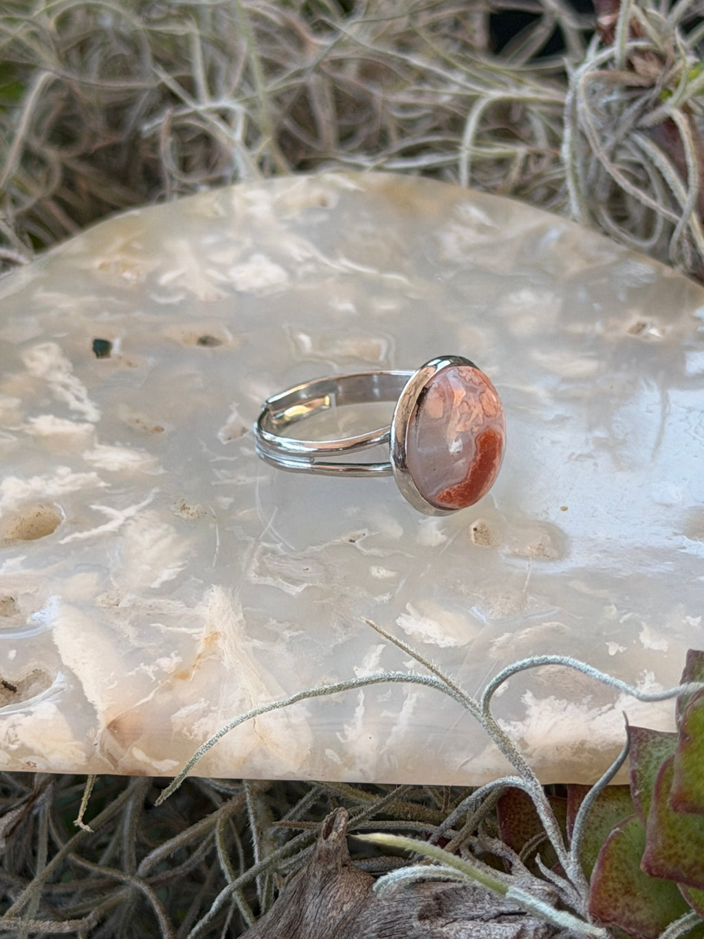 A silver ring with a round, peachy-orange stone set on a polished white agate slab surrounded by dried grass and small green leaves.