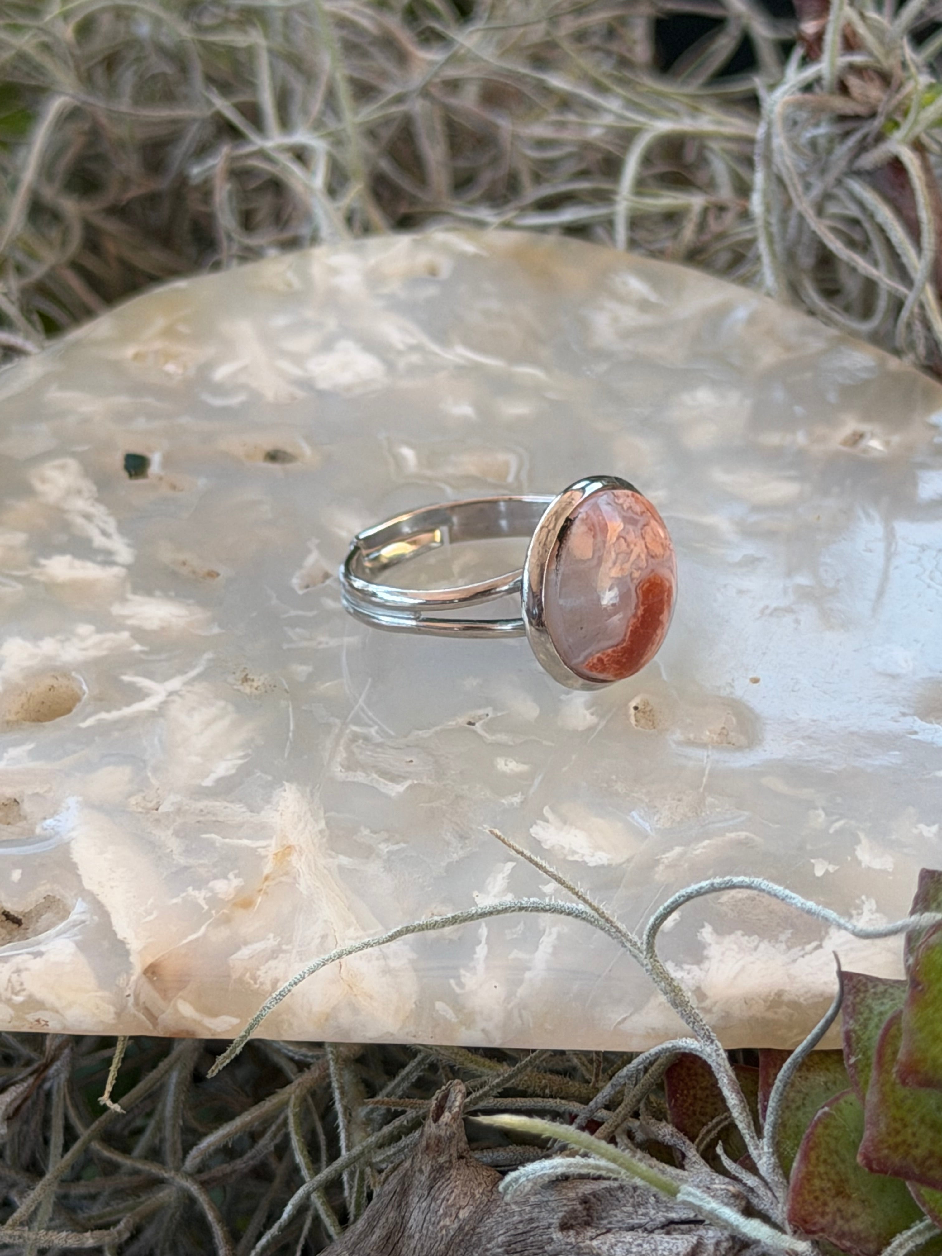 A silver ring with a round, peachy-orange stone set on a polished white agate slab surrounded by dried grass and small green leaves.