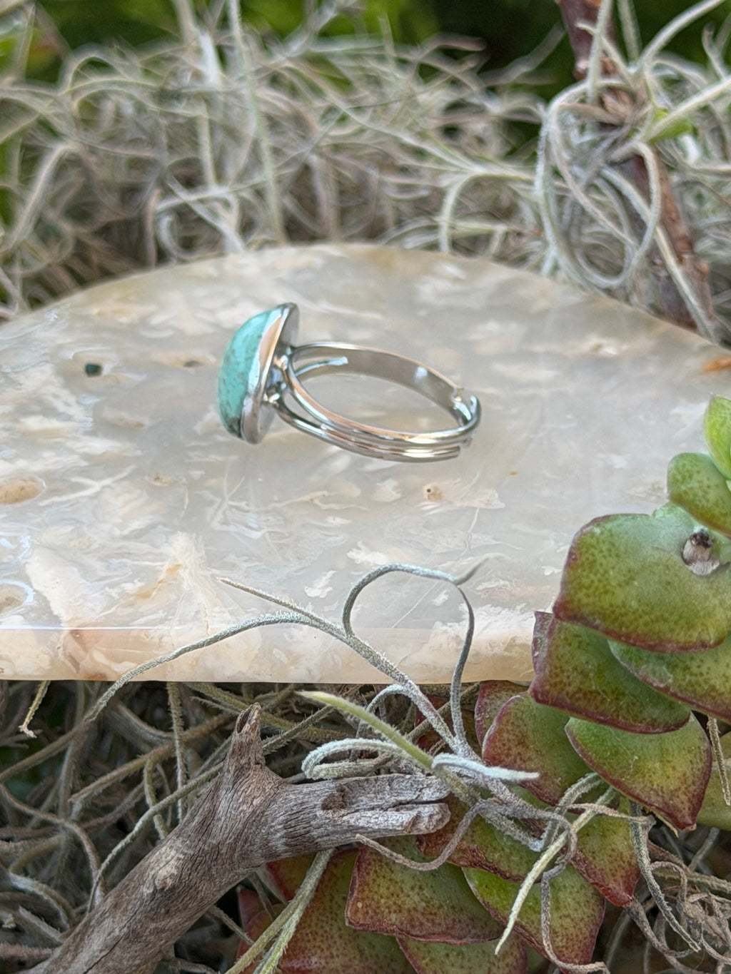A silver ring with a small oval light blue-green stone, displayed on a textured white stone slab surrounded by dried grass and succulent leaves.