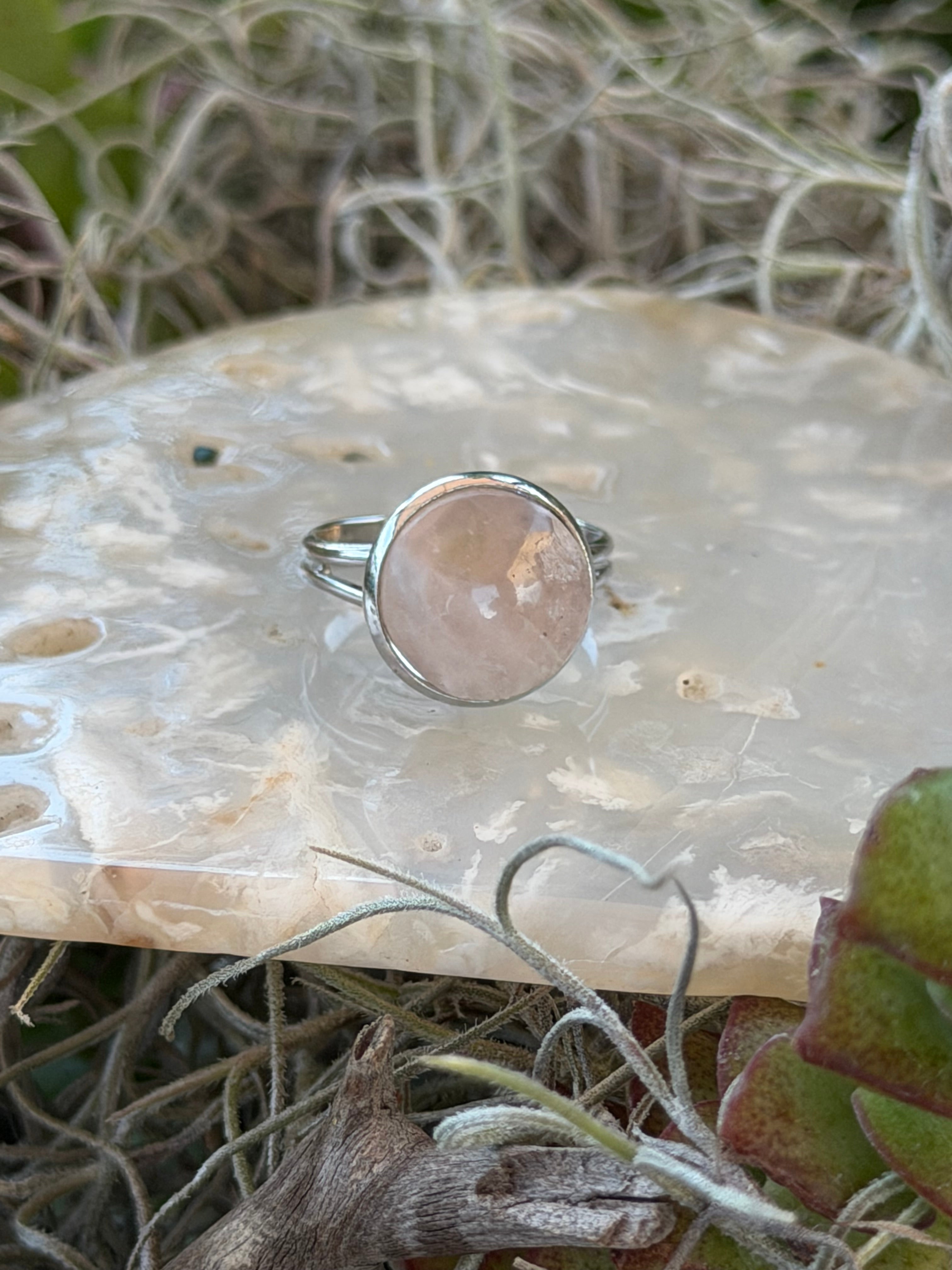 A silver ring with a round, polished pale pink stone, displayed on a textured white stone slab surrounded by dried grass and green leaves.