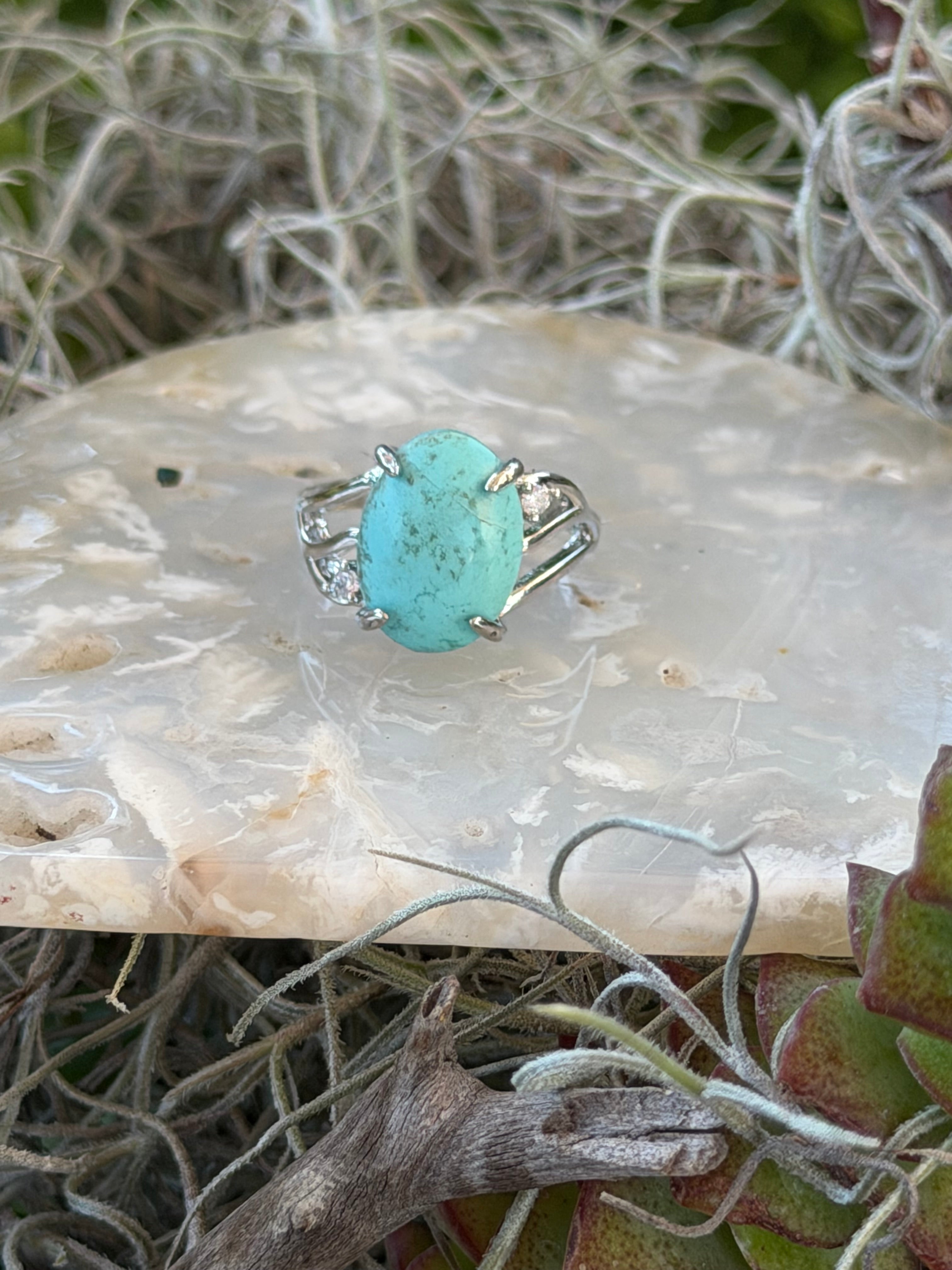 A silver ring featuring a round turquoise cabochon stone, displayed on a piece of white agate with dried grass and succulent foliage in the background.