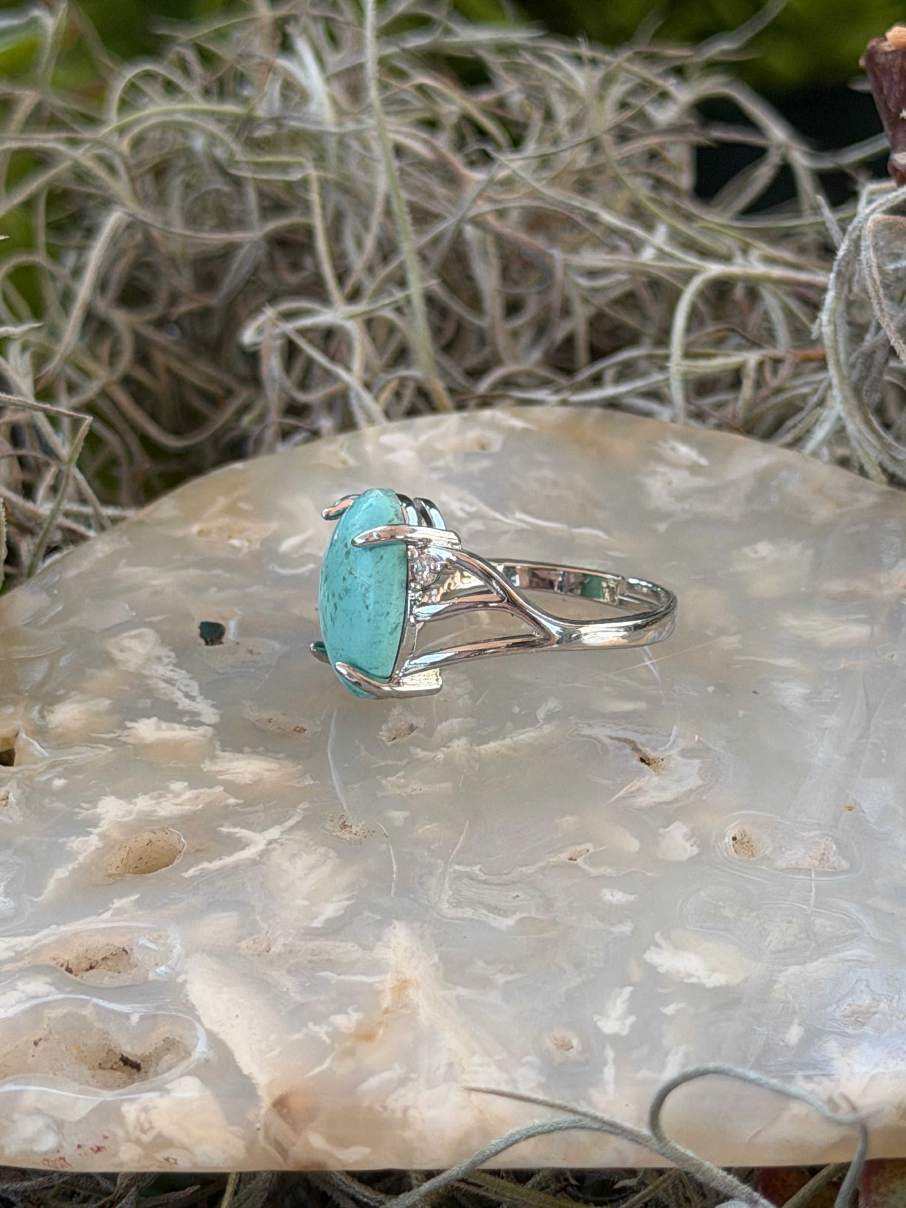 A silver ring featuring a large oval turquoise stone, displayed on a polished agate slab with dried moss as a decorative background.