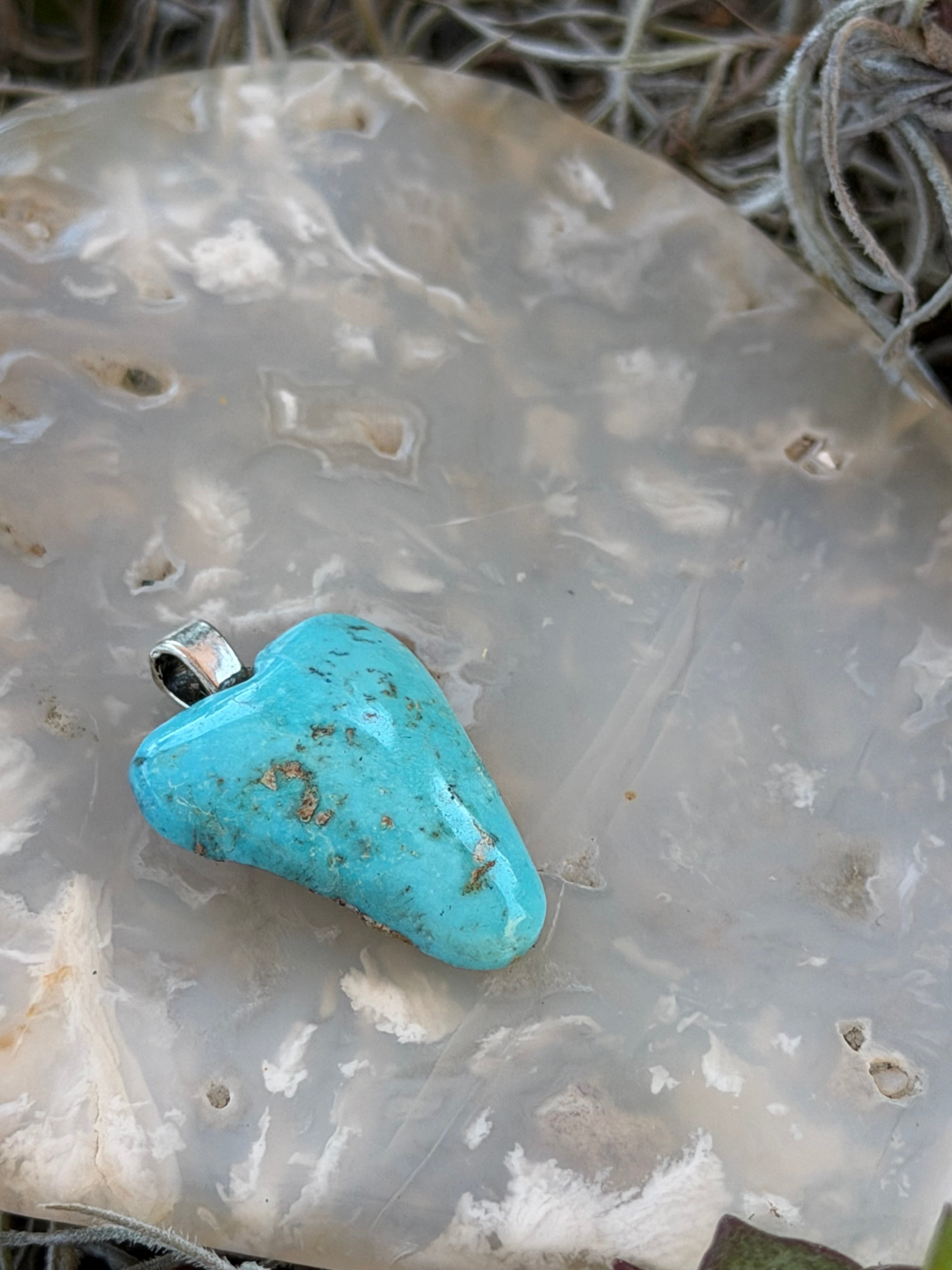 A small turquoise stone pendant with a silver bail, resting on a textured, translucent white agate slab.