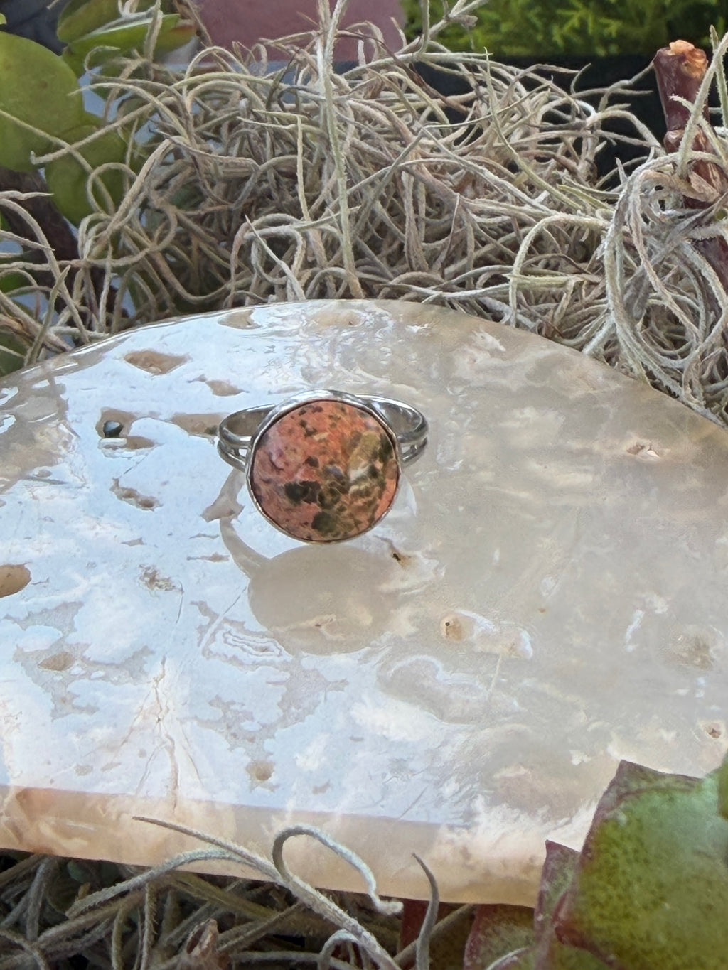 A silver ring with a round, mottled orange-brown stone, displayed on a polished white stone slab surrounded by dried grass and green leaves.