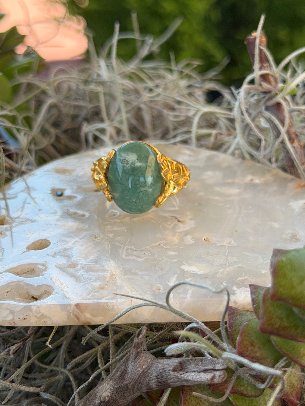 A gold ring with a green oval stone, displayed on a white stone slab surrounded by dried grass and green foliage.