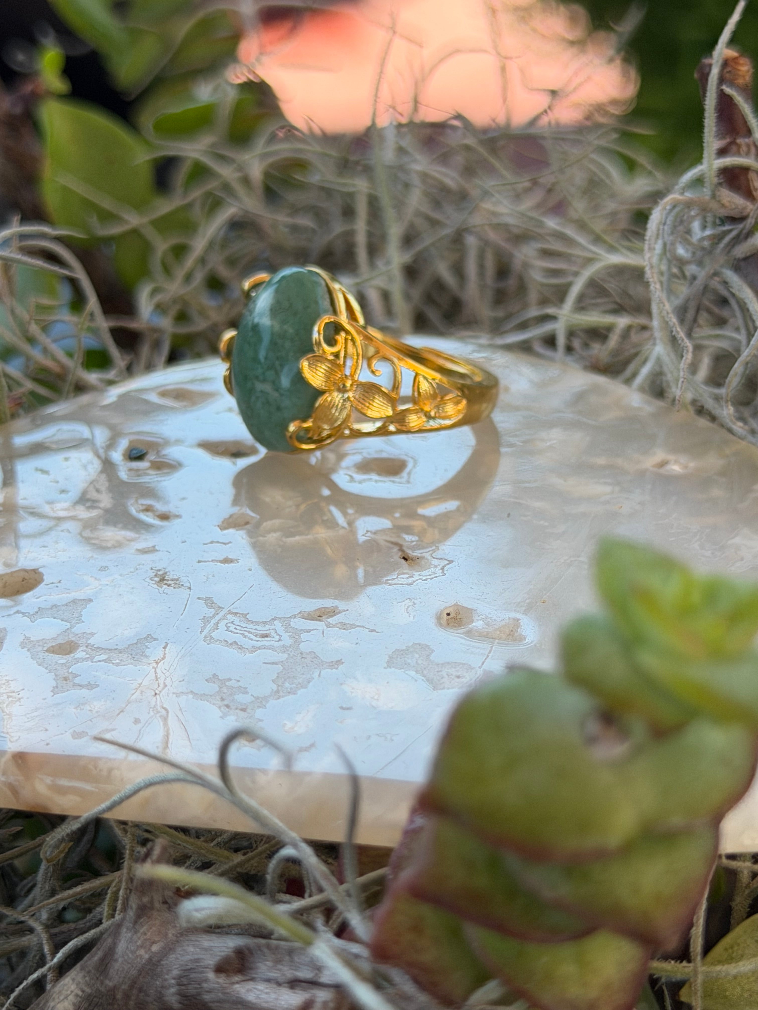 A gold ring with a green stone, photographed close-up on a white stone slab surrounded by dried grass and green leaves.