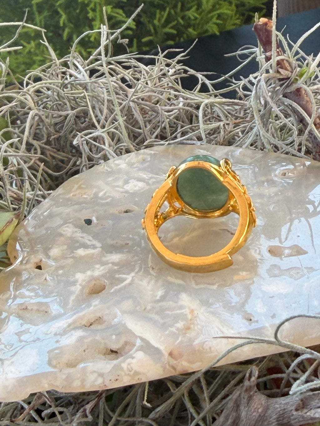 A gold ring with a green teardrop-shaped stone, displayed on a polished white crystal cluster surrounded by dried grass.