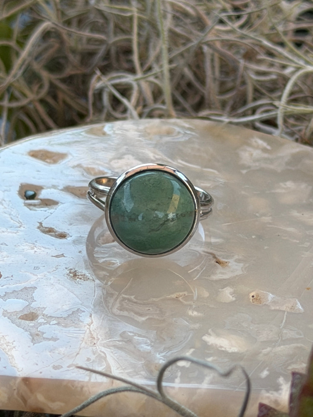 A silver ring featuring a round, polished green stone, displayed on a textured white stone with dried grass in the background.