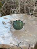 A silver ring featuring a round, polished green stone, displayed on a textured white stone with dried grass in the background.