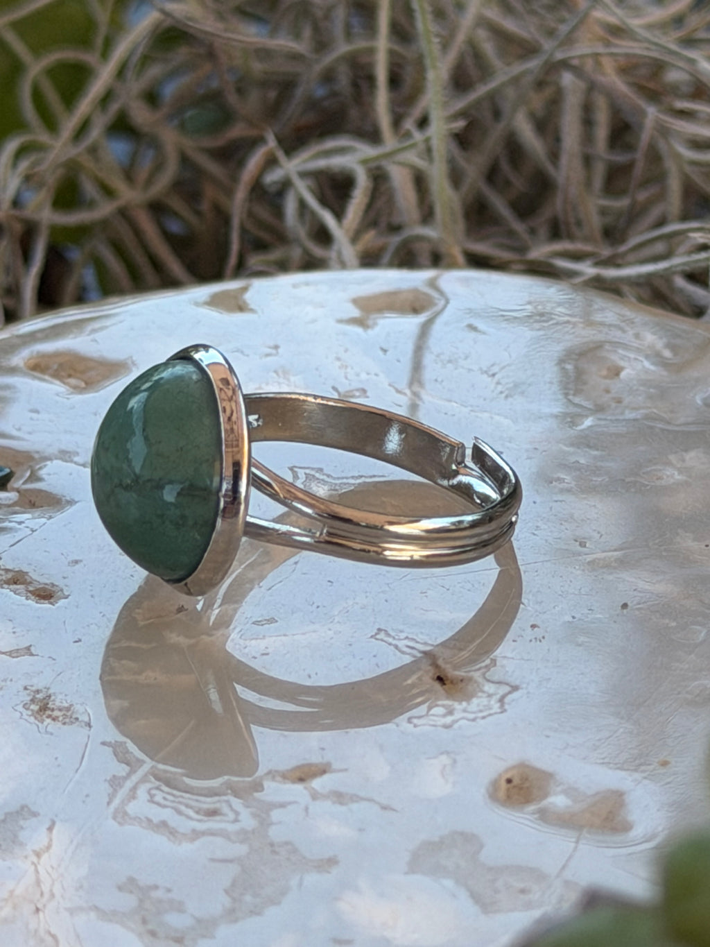 A silver-toned ring with a polished green stone, displayed on a textured white surface with natural twigs in the background.