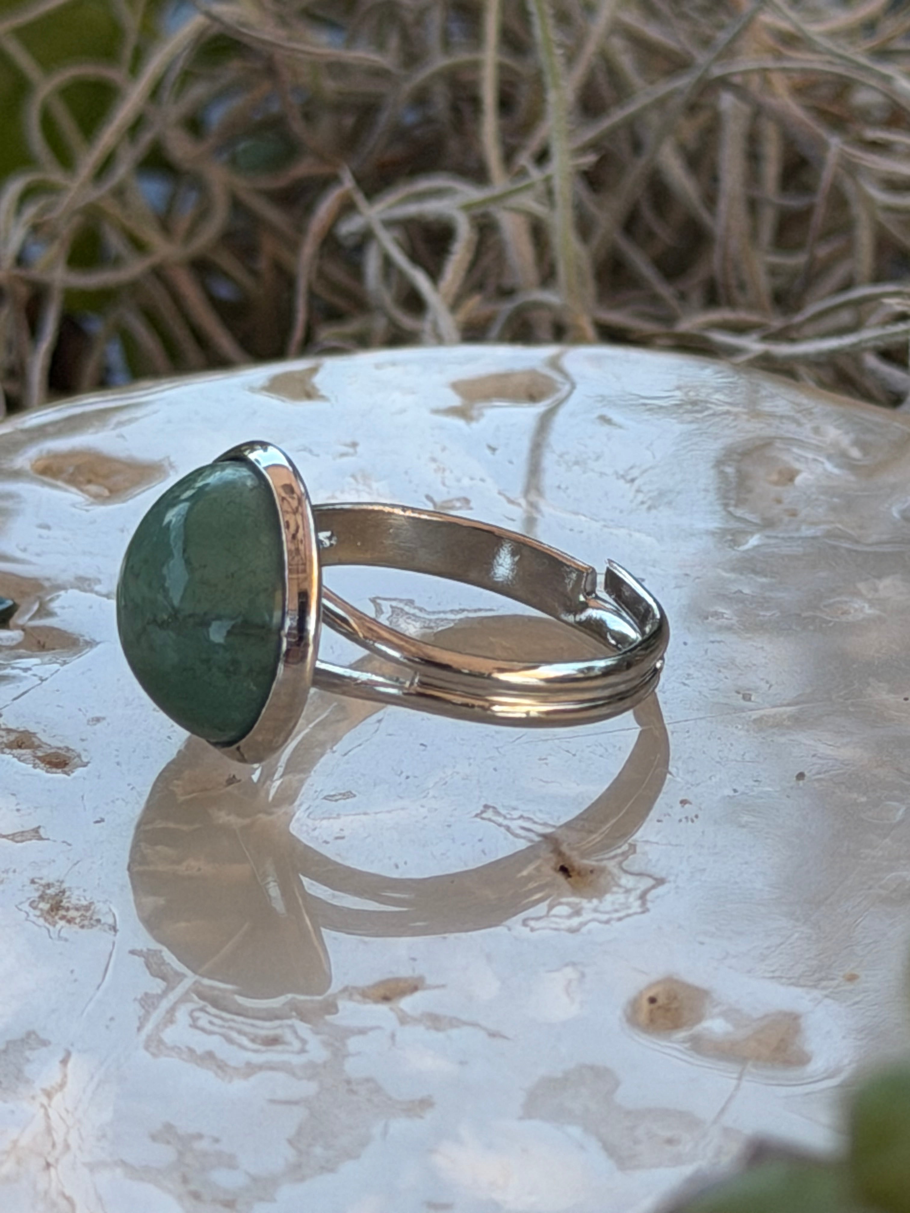 A silver-toned ring with a polished green stone, displayed on a textured white surface with natural twigs in the background.