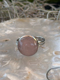 A silver ring featuring a large, round, dusty pink rose quartz cabochon, displayed on a textured white stone surface with dried grass in the background.