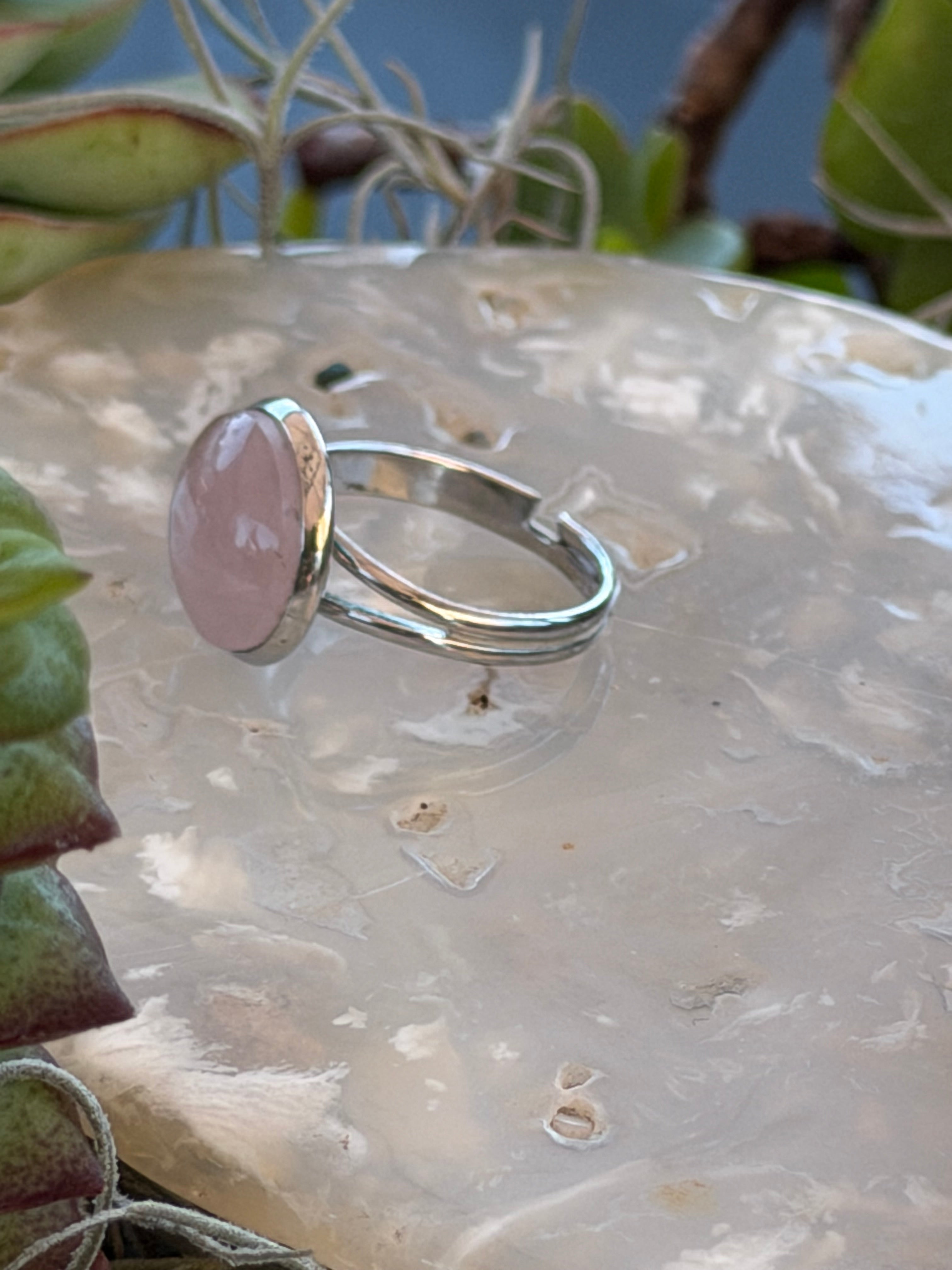 A silver ring with a large, oval, light pink stone, displayed on a textured, translucent stone slab with succulents in the background.