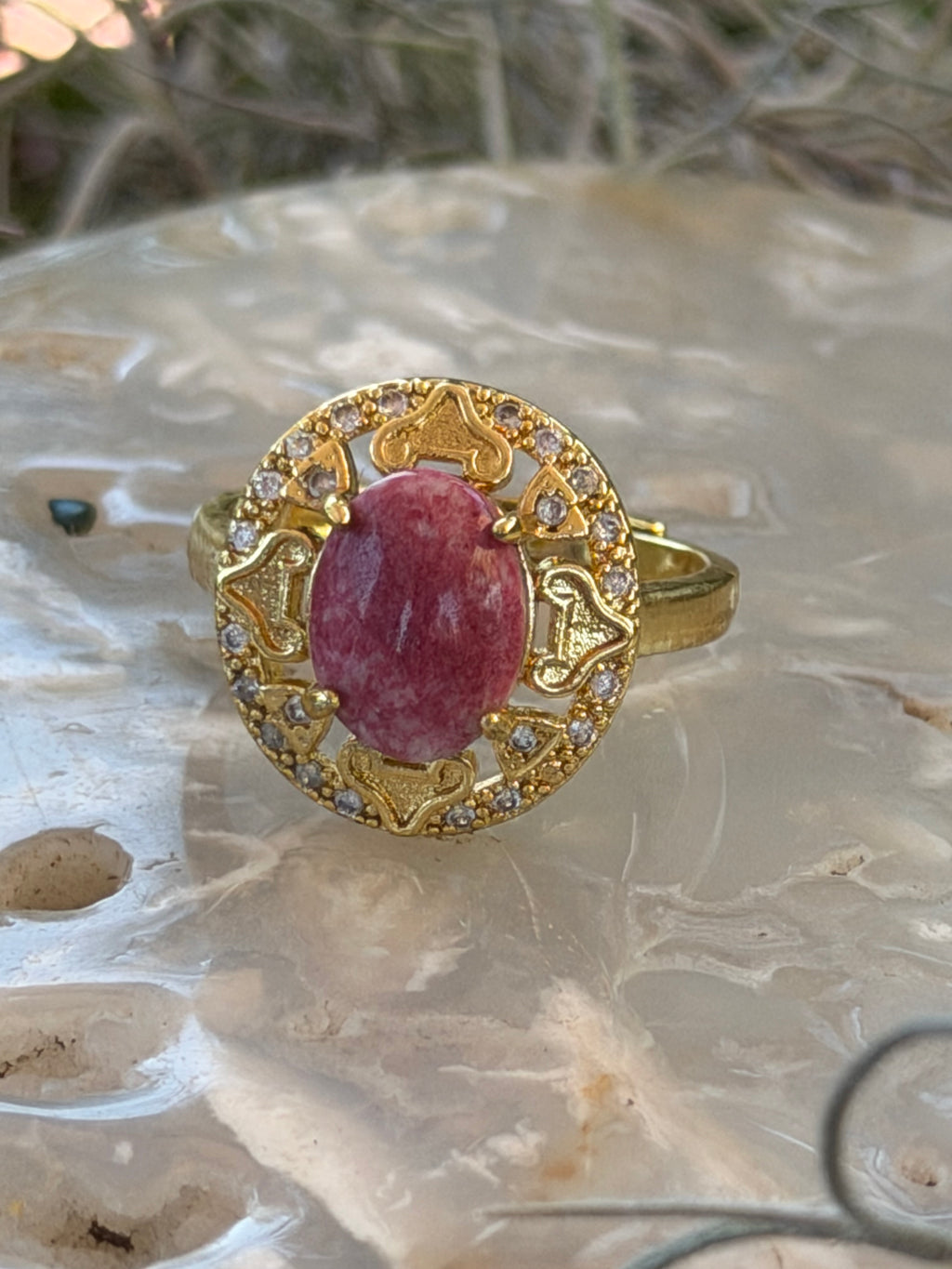 A gold ring with an oval pink gemstone and small clear stones, displayed on a textured white shell.