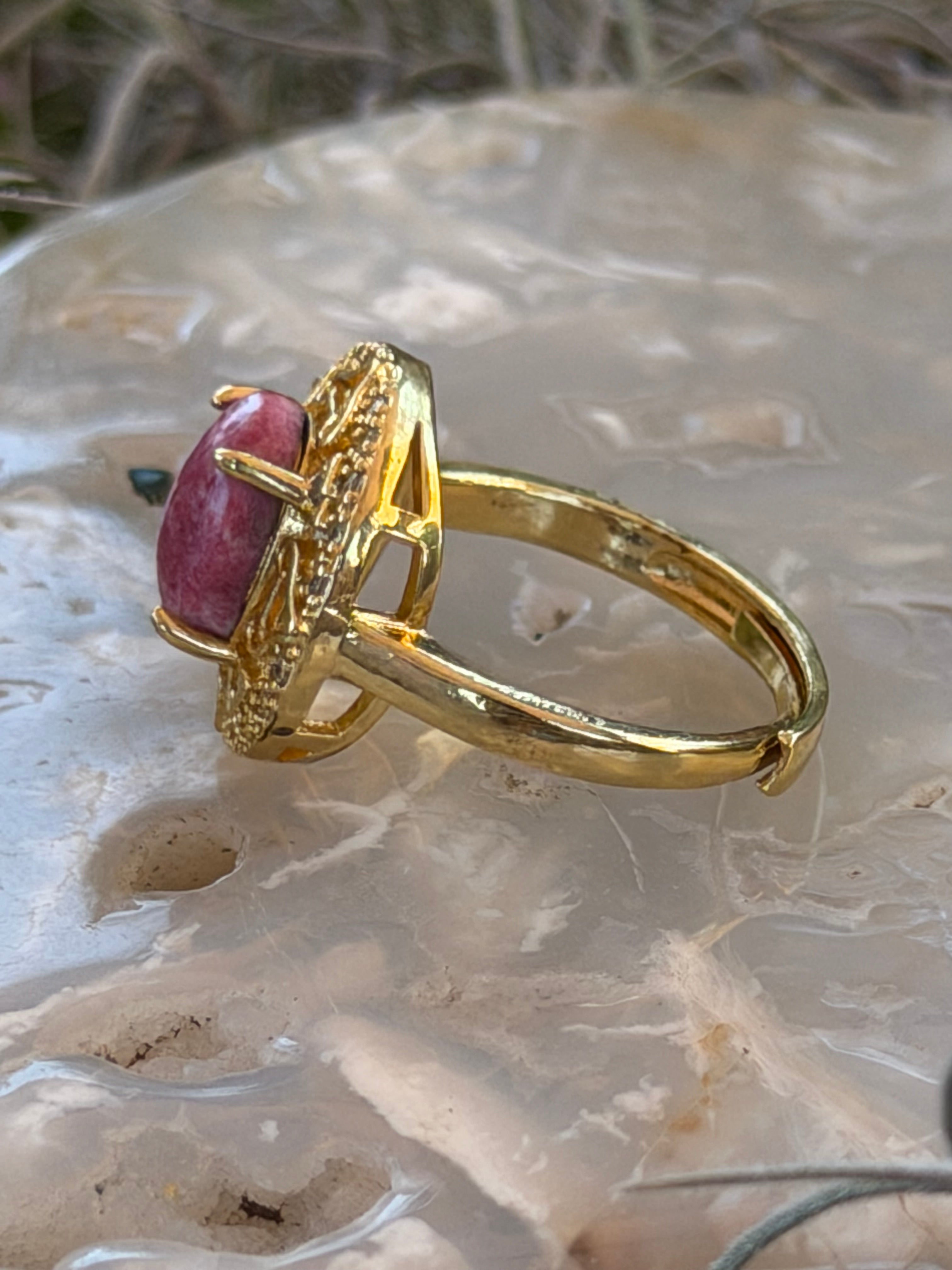 A gold hoop-style ring with a decorative openwork band and a pinkish-red oval gemstone, photographed close-up against a translucent white stone surface.