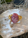 A gold-toned jewelry piece featuring an oval pinkish-red stone with white marbling, displayed on a polished white agate slab with dried grass.