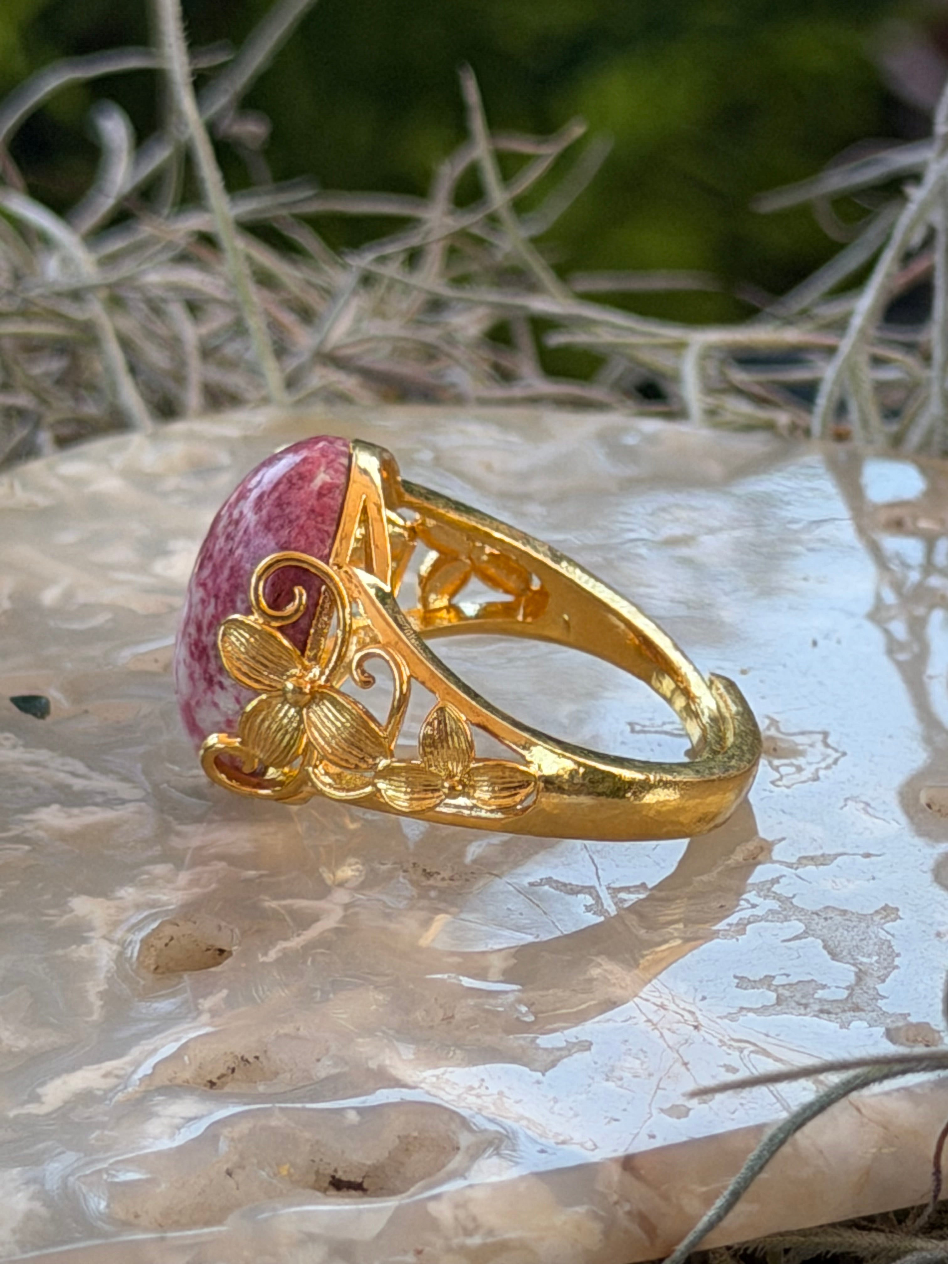 A gold filigree ring with a pink and white marbled cabochon stone, displayed on a textured white surface with blurred green foliage in the background.