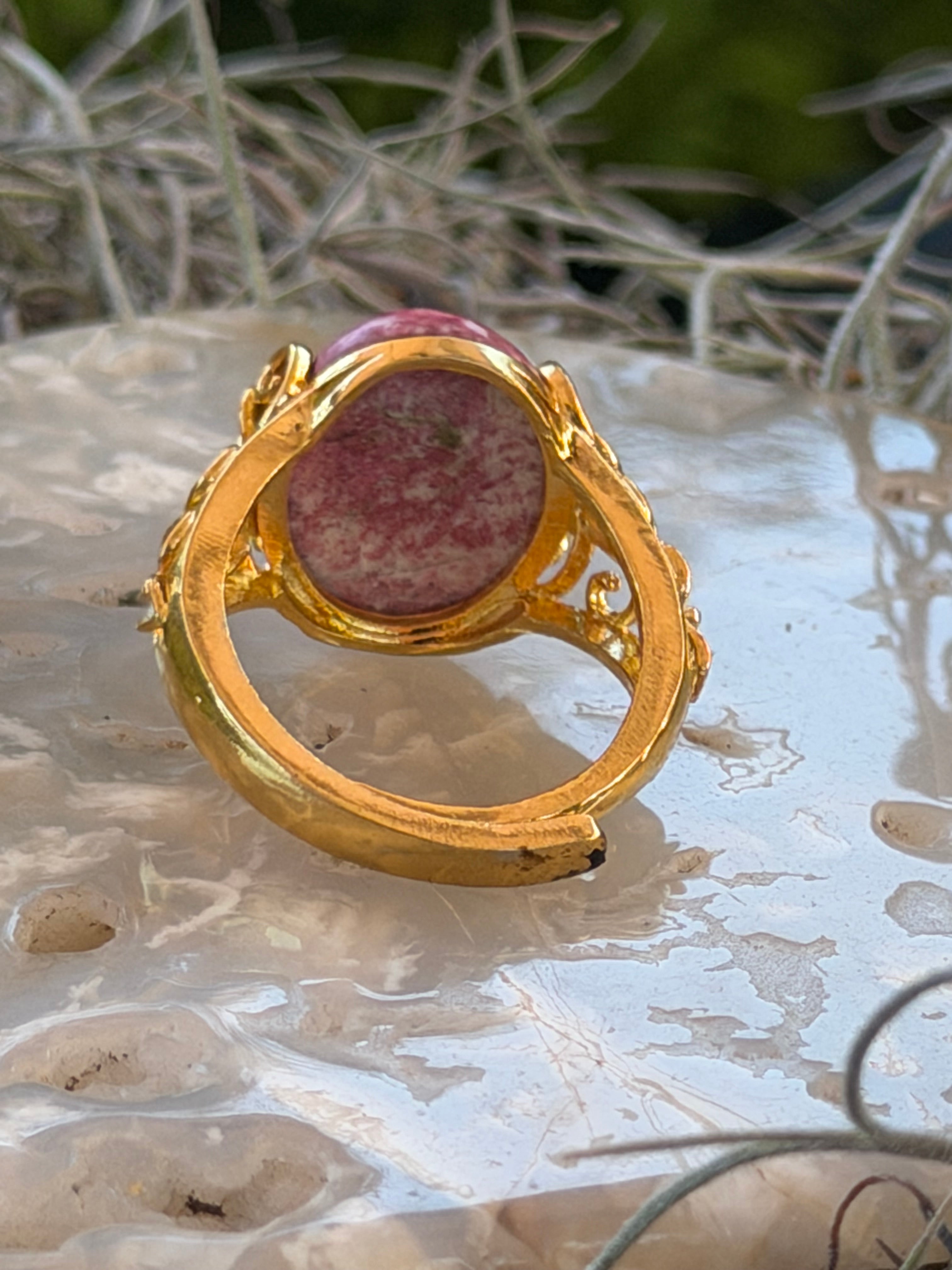 A gold ring with a round pinkish-red stone, photographed close-up against a textured white surface with some blurred natural elements in the background.
