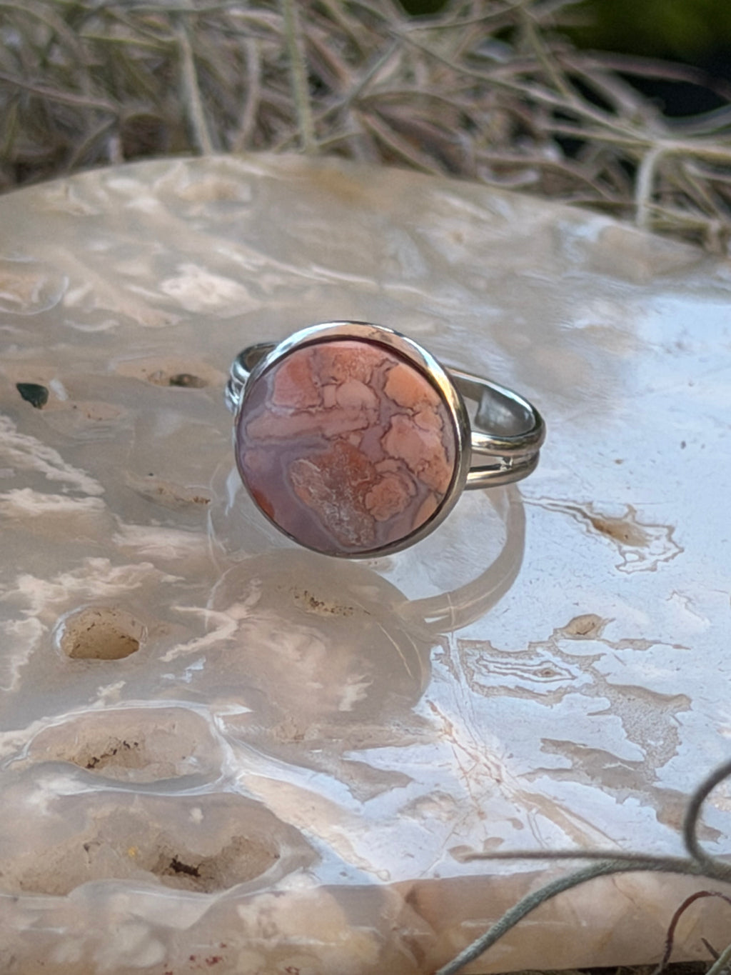 A silver band ring featuring a round cabochon stone with a pinkish-brown and orange marbled pattern, displayed on a textured white surface.