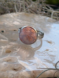 A silver band ring featuring a round cabochon stone with a pinkish-brown and orange marbled pattern, displayed on a textured white surface.