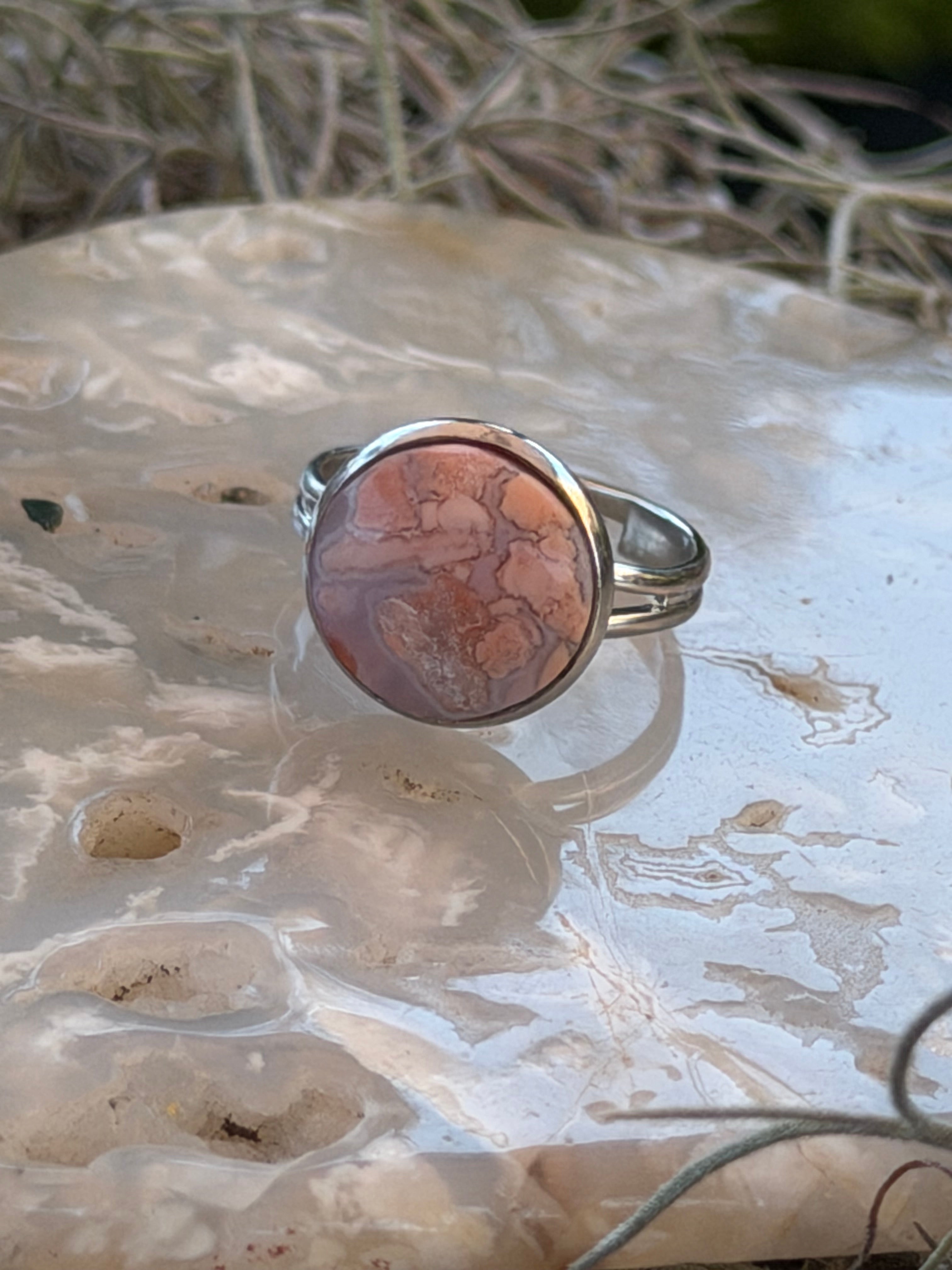 A silver band ring featuring a round cabochon stone with a pinkish-brown and orange marbled pattern, displayed on a textured white surface.