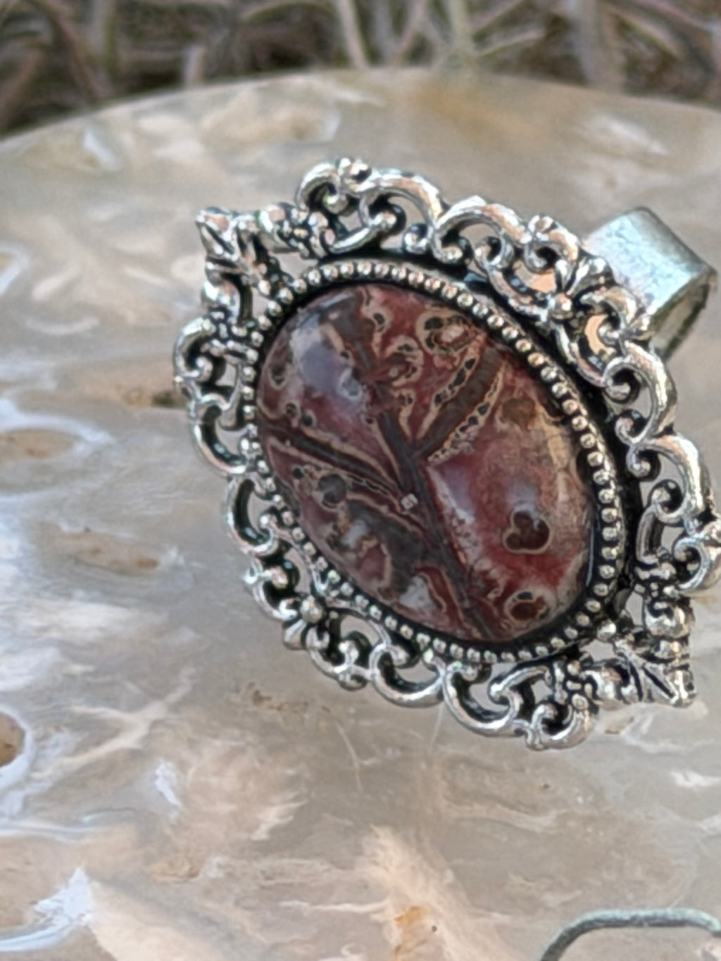 An ornate silver-toned ring featuring a round cabochon gemstone with a brown and reddish marbled pattern, displayed on a textured light stone surface.