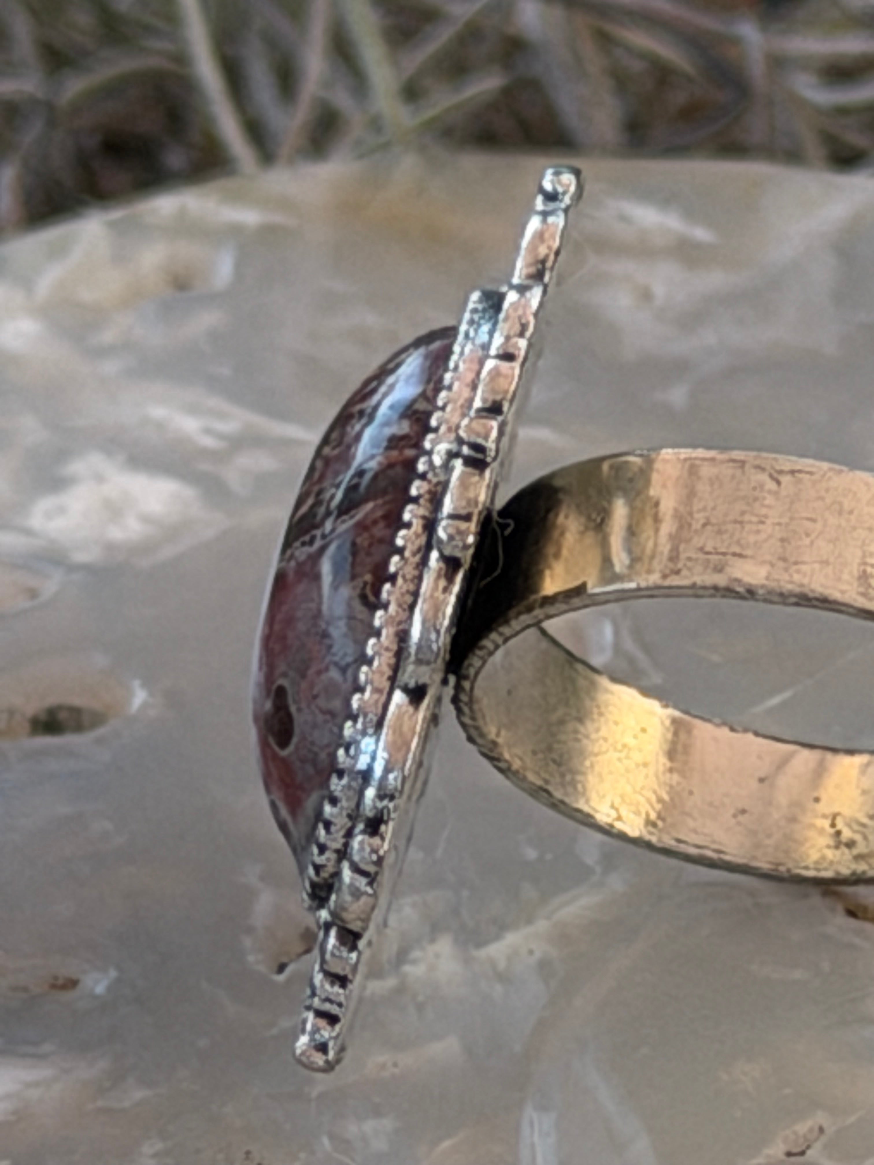 A close-up of a handmade ring featuring a polished gold band and a silver-toned bezel-set oval cabochon with a dark reddish-brown marbled inlay, resting on a translucent gray stone.