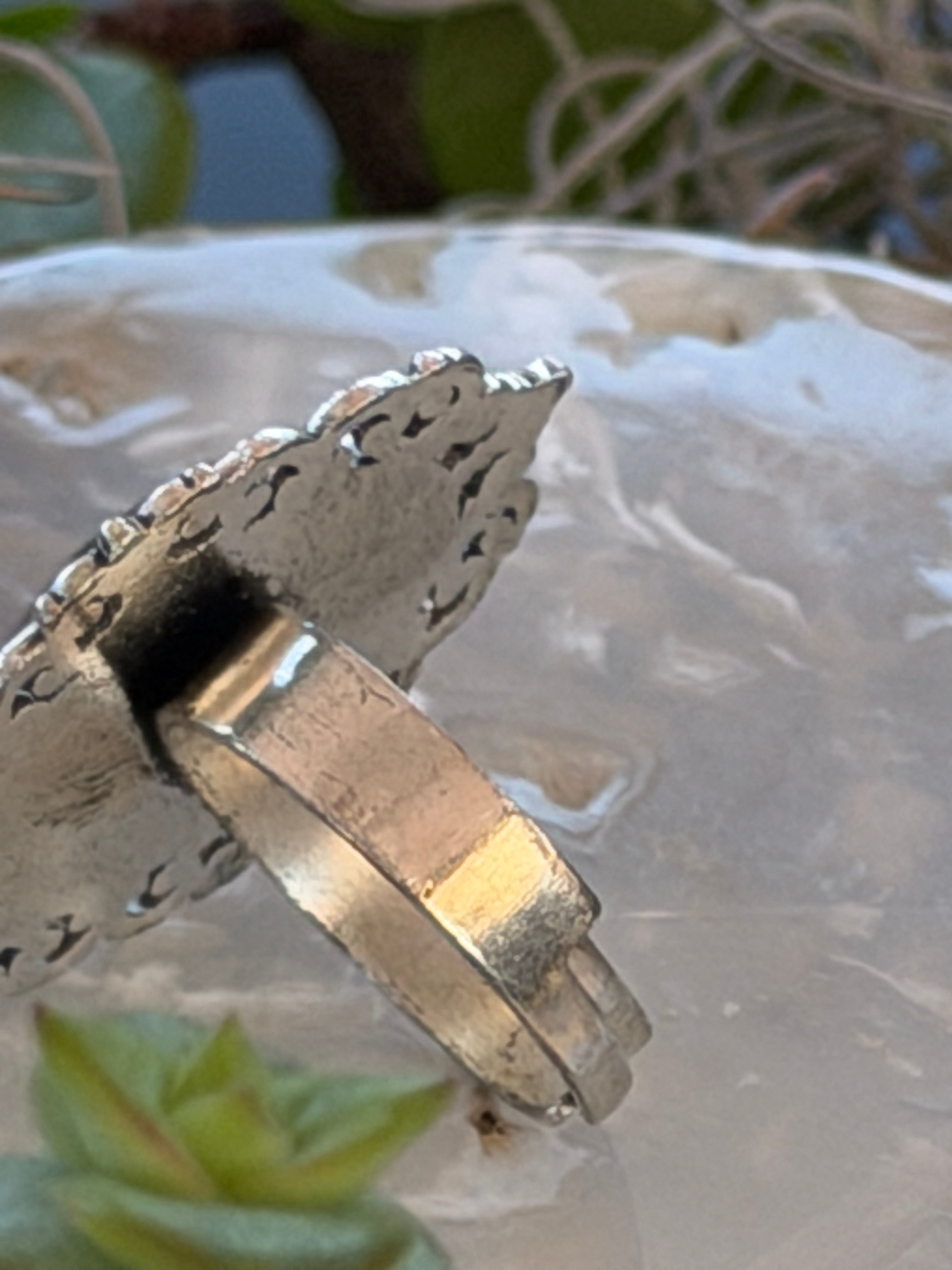 A close-up of a silver-toned metal ring with a decorative, textured band, shown against a translucent white glass background with blurred green foliage.