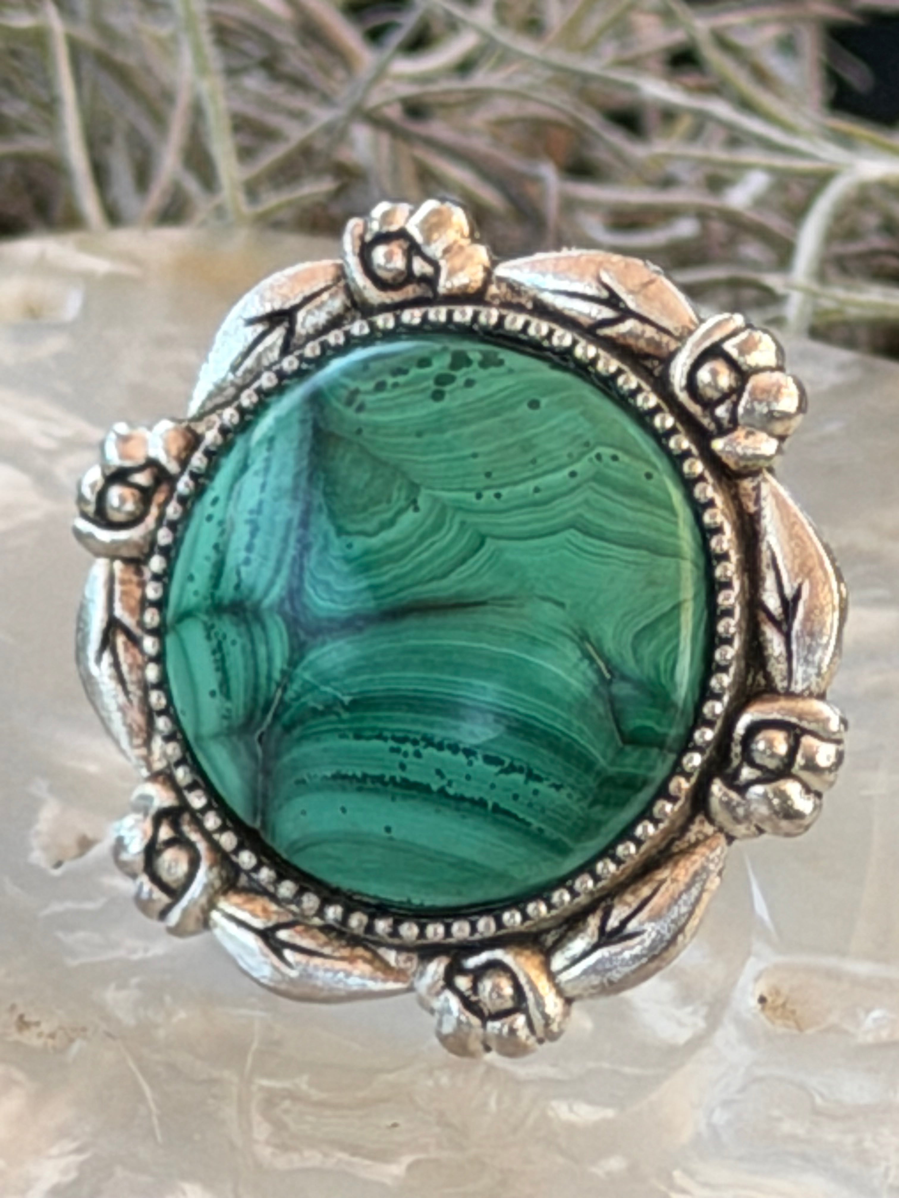 A close-up of a round green malachite stone set in an ornate silver-toned floral bezel, resting on a pale stone surface with blurred dried grass in the background.