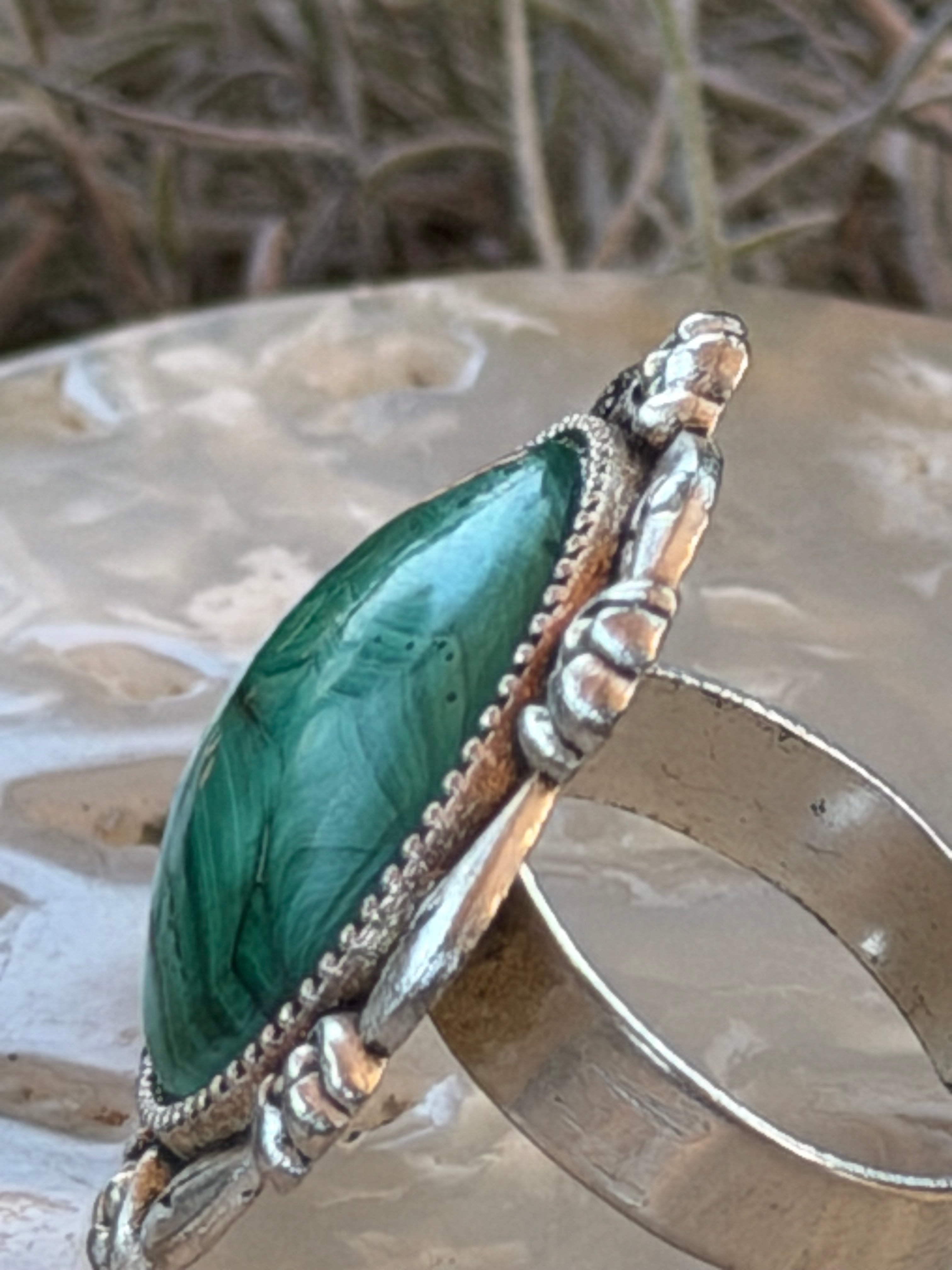 A close-up of a silver-toned ring featuring a large, polished Malachite with natural marbled patterns, displayed on a textured metallic surface.